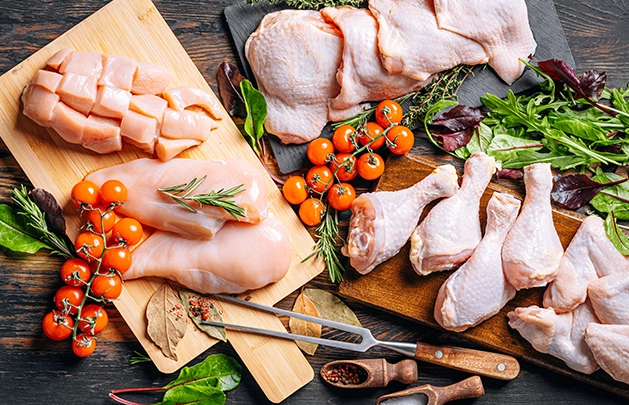 eagle eyes view of a wooden table that has three cutting boards on it with different cuts of chicken. One has thighs with herbs next to it, one has drumsticks with lettuce and tomatoes next to it, and the other has freshly diced and prepared chicken breasts with tomatoes, salt and pepper and a carving fork next to it