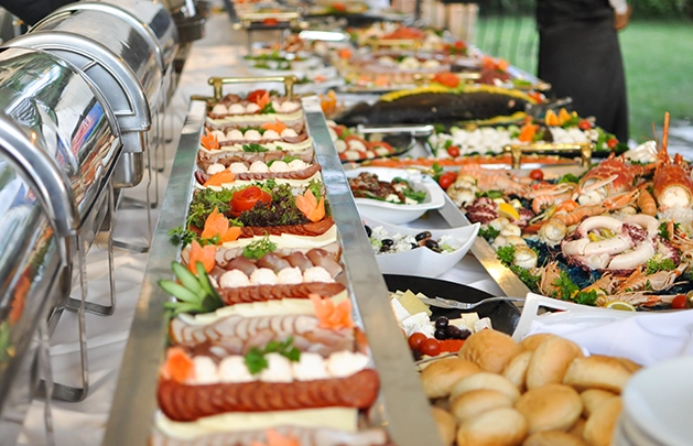 side view of a catering spread that has hot prepared foods in silver casings, charcuterie boards, seafood platters, rolls, and entrees