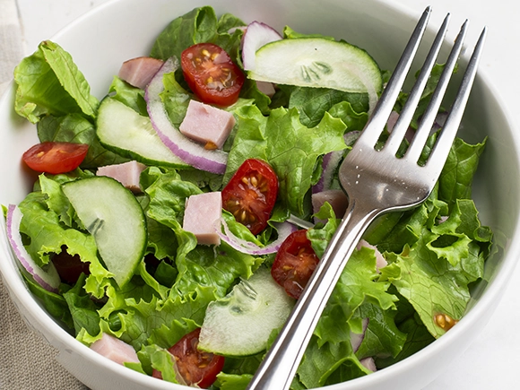 bowl of green salad with tomatoes, cut up red onions, ham and cucumbers in the bowl with a fork placed on to of the bowl.